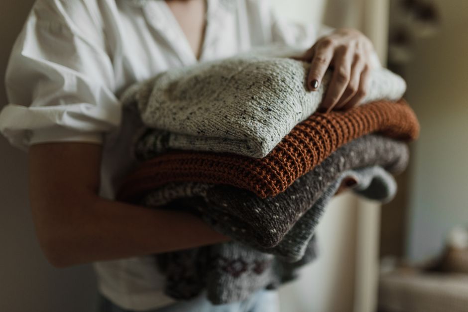 A woman holds a stack of folded knit sweaters in warm autumn colors indoors.