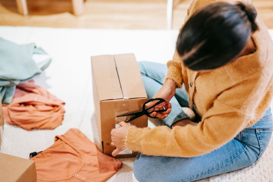 Crop anonymous female using scissors while packing carton box with parcel on floor among casual clothes